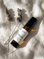 A bottle of kiln Dusk essential oil roll-on, placed diagonally on a textured beige linen fabric alongside two dried lavender sprigs under warm, direct sunlight.
