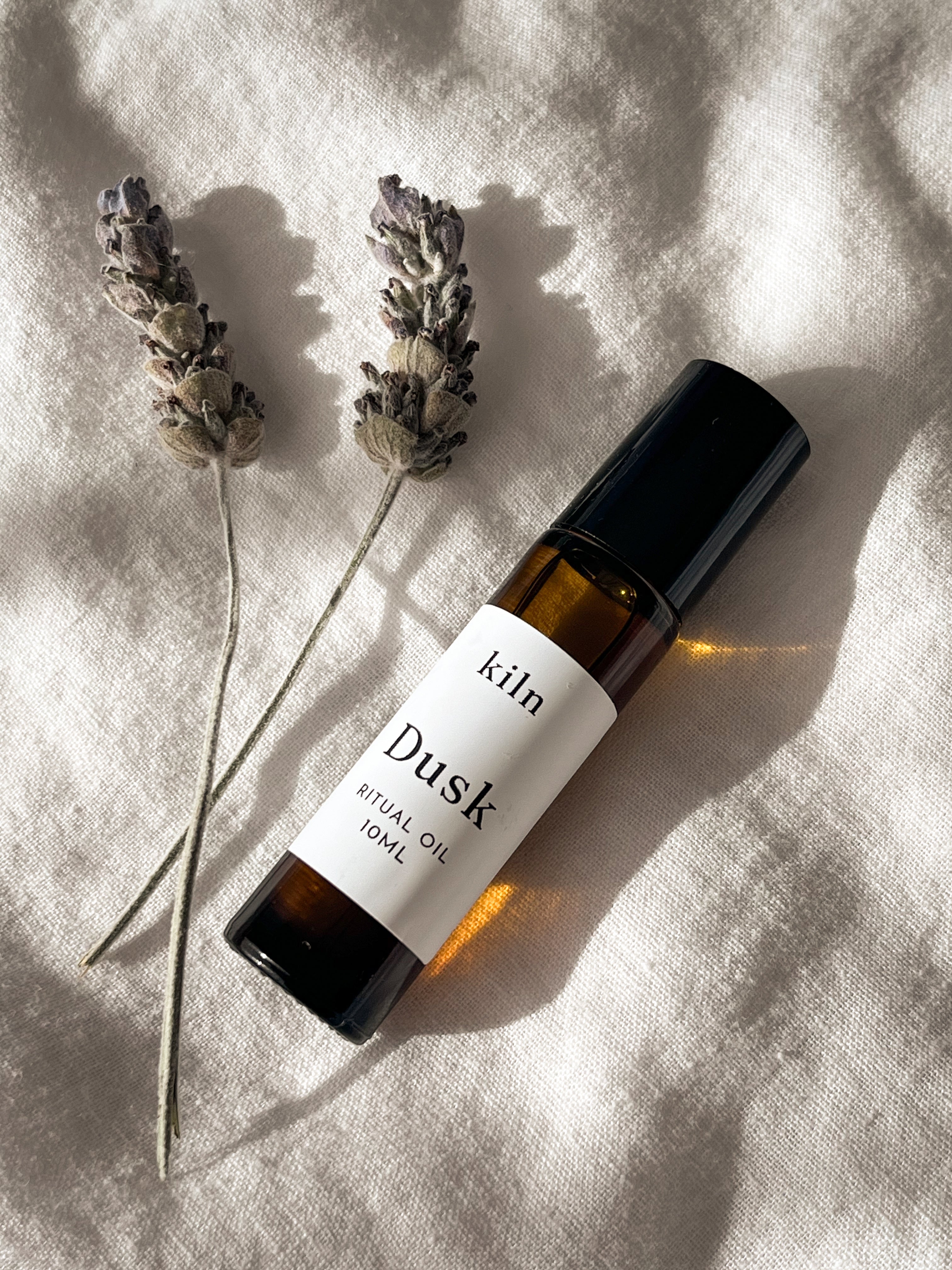 A bottle of kiln Dusk essential oil roll-on, placed diagonally on a textured beige linen fabric alongside two dried lavender sprigs under warm, direct sunlight.
