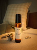 A 10ml bottle of kiln Dusk Ritual Oil stands on a light beige tabletop in a cozy bedroom setting, with a checkered pillow in the background and dried herbs and rings as props.