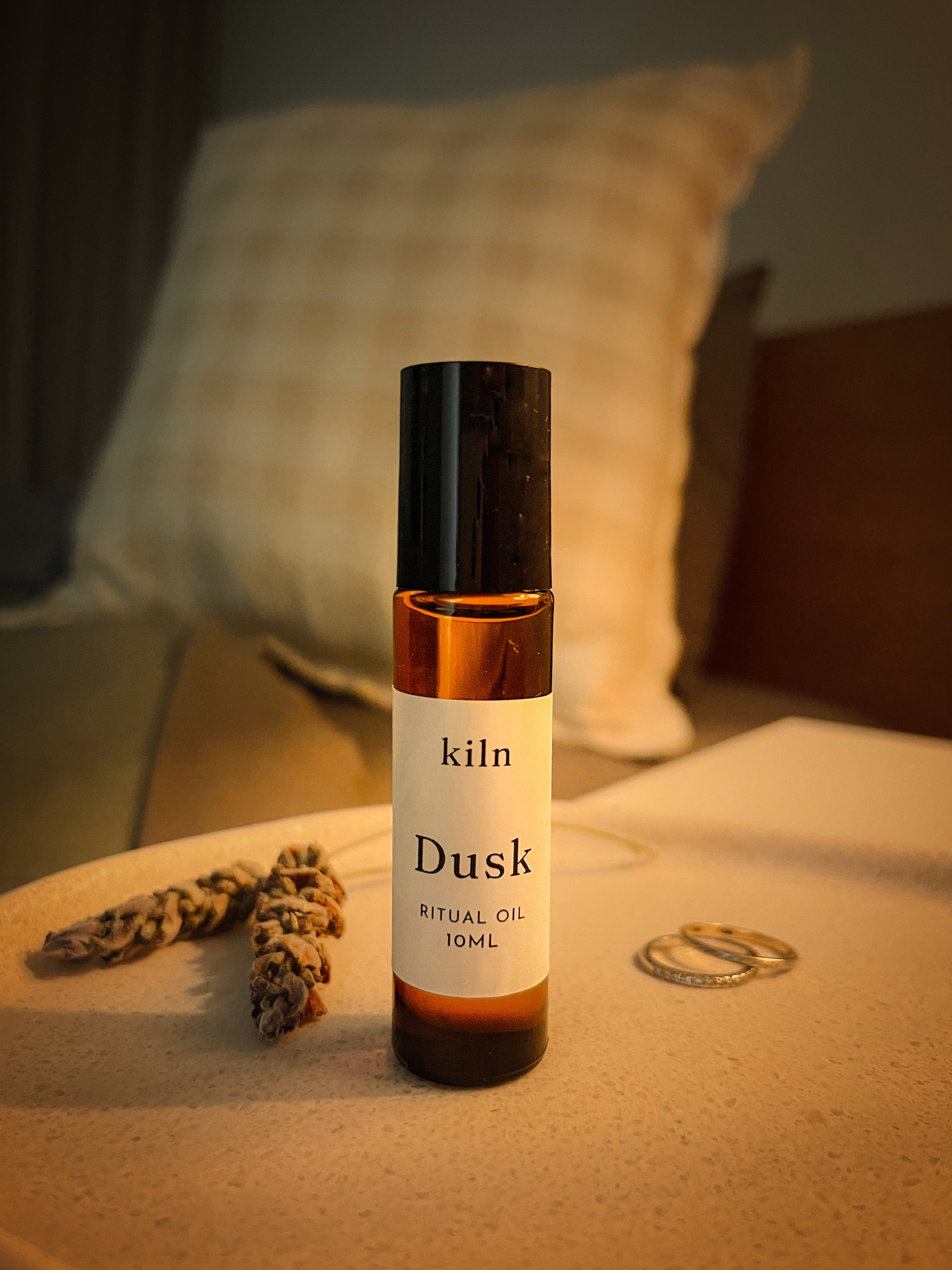 A 10ml bottle of kiln Dusk Ritual Oil stands on a light beige tabletop in a cozy bedroom setting, with a checkered pillow in the background and dried herbs and rings as props.