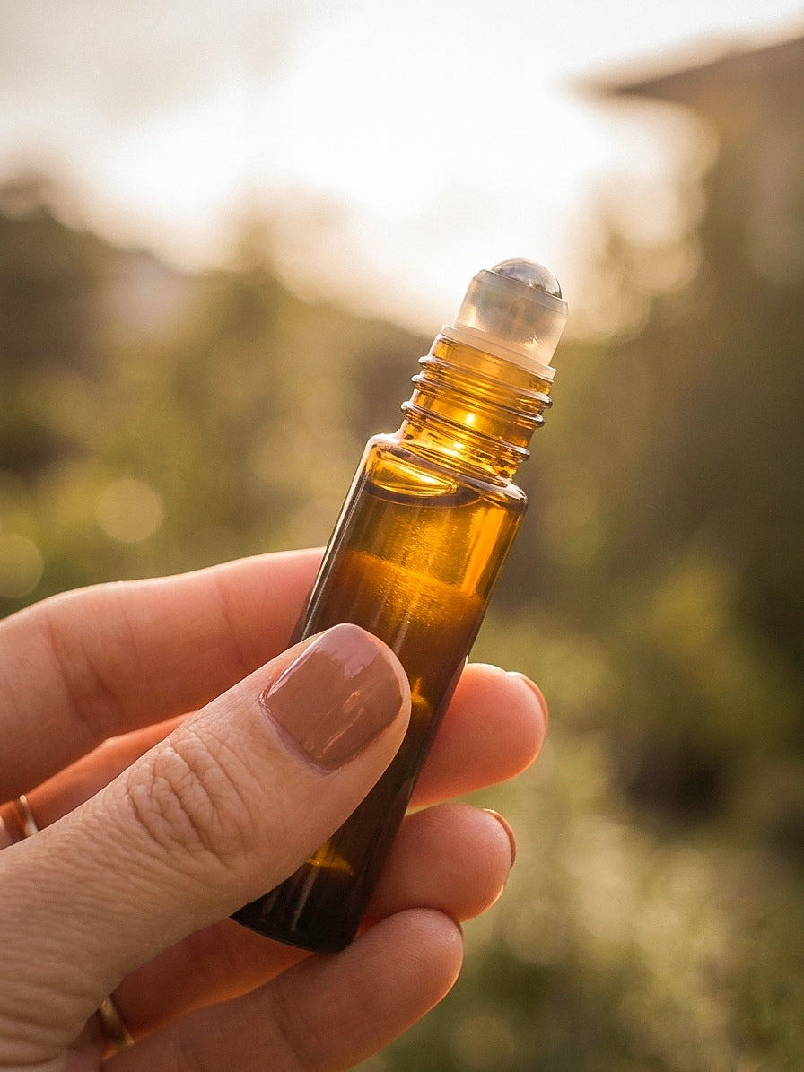 Hand holding a small amber glass perfume oil roller bottle with a blurred natural background